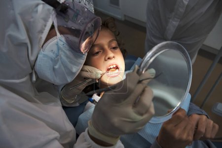 Foto de Alto ángulo del dentista anónimo en traje protector examinando la boca del paciente con herramientas en la clínica durante el brote de coronavirus - Imagen libre de derechos