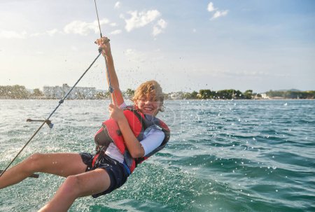 Téléchargez les photos : Goutte d'eau éclaboussures sur ravi garçon préadolescent aux cheveux blonds dans la vie gilet équilibrage sur corde sur la mer ondulée pendant les vacances d'été actives - en image libre de droit