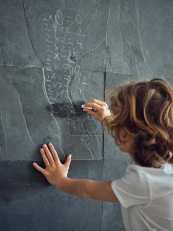 Téléchargez les photos : Vue arrière d'un enfant intelligent résolvant des équations mathématiques sur un mur de pierre noire avec un morceau de craie - en image libre de droit