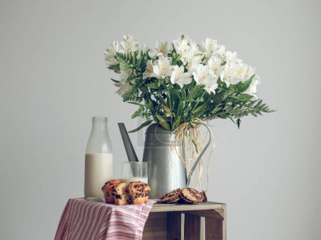 Foto de Delicadas flores blancas en regadera se colocan cerca de deliciosos pasteles y botellas de leche en la caja de madera en el estudio - Imagen libre de derechos