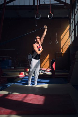 Téléchargez les photos : Jeune gymnaste sportif s'étirant avec un élastique dans le gymnase avant de faire ses exercices. - en image libre de droit