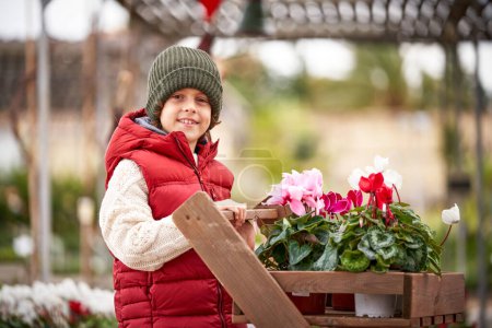 Photo for Cute boy in knitted hat standing near wooden cart with vivid blossoming flowers in pots in botanic garden - Royalty Free Image