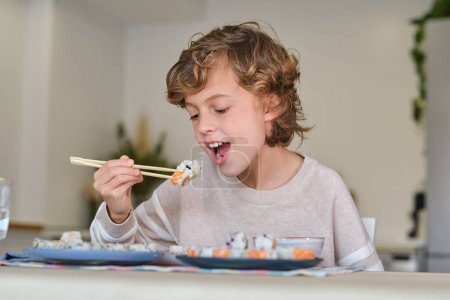 Téléchargez les photos : Mignon enfant avec baguettes ouverture bouche tandis que assis à la table dans la cuisine et manger savoureux sushi - en image libre de droit