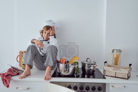 Foto de Cuerpo completo de lindo niño preadolescente en gorra de chef y traje sentado en el mostrador de la cocina cerca de la estufa con cacerola con zanahorias y espaguetis - Imagen libre de derechos