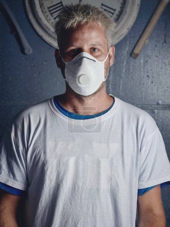 Photo for Serious adult ethnic blond haired make shaper with tattoos wearing protective respirator mask and white t shirt standing near wall with clock and looking at camera - Royalty Free Image