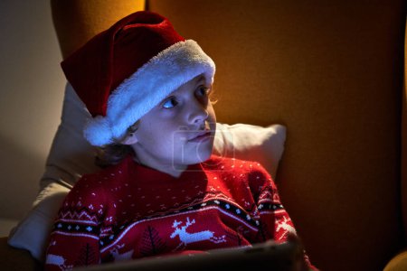 Téléchargez les photos : Charmant garçon en pull chaud et chapeau de Père Noël avec tablette relaxante dans un fauteuil doux tout en regardant loin dans la pièce sombre avec lampe - en image libre de droit