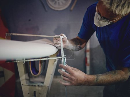 Photo for Crop attentive male shaper with blond hair and tattoos on arms in casual clothes and respirator measuring surfboard thickness with caliper during work in joinery - Royalty Free Image