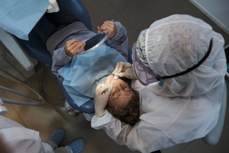 Téléchargez les photos : Vue du dessus d'un dentiste méconnaissable en costume de protection examinant les dents d'un enfant allongé sur une chaise dentaire pendant une pandémie de coronavirus - en image libre de droit