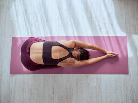 Téléchargez les photos : Vue du dessus corps entier de femme active anonyme faisant la posture Utthita Balasana sur tapis rose pendant la séance de yoga dans la chambre - en image libre de droit