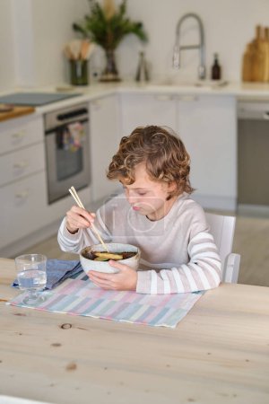 Téléchargez les photos : Focalisé enfant assis à la table en bois et manger de la nourriture asiatique avec des baguettes dans la cuisine confortable à la maison - en image libre de droit