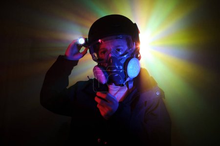 Photo for Boy in gas mask with portable walkie talkie in hand shining bright flashlight while robbing dark mint with colorful neon lights - Royalty Free Image