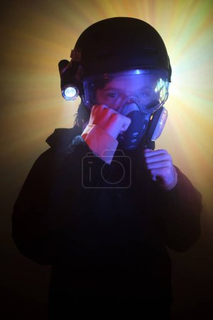 Téléchargez les photos : Enfant sérieux dans un masque à gaz protecteur avec une lampe de poche brillante regardant la caméra avec des poings serrés prêts pour le combat tout en cambriolant la menthe sombre contre l'éclairage au néon coloré - en image libre de droit