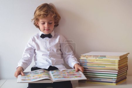 Téléchargez les photos : Garçon heureux en tenue formelle avec noeud papillon lecture livre intéressant tout en étant assis sur la table près de fond blanc dans la chambre - en image libre de droit