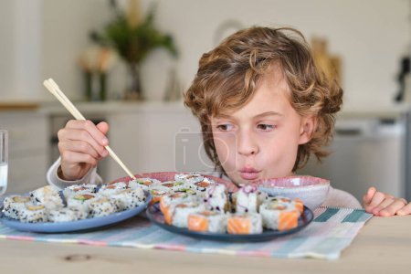 Téléchargez les photos : Enfant curieux assis à table et touchant savoureux rouleaux de sushi avec baguettes en bois tout en déjeunant dans la cuisine à la maison - en image libre de droit