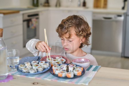 Téléchargez les photos : Enfant curieux aux cheveux bouclés assis à table et regardant vers le bas avec intérêt tout en mangeant de savoureux rouleaux de sushi avec des baguettes dans la cuisine à la maison - en image libre de droit