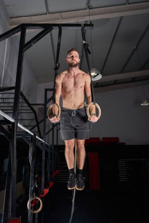 Foto de Cuerpo completo de deportista muscular con torso desnudo colgado en anillos gimnásticos durante el entrenamiento funcional en luz tenue - Imagen libre de derechos