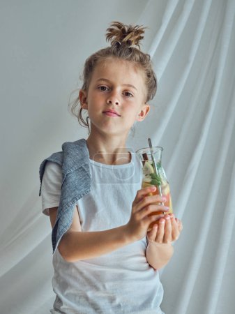 Téléchargez les photos : Enfant avec coiffure drôle tenant verre de limonade maison rafraîchissante faite de divers agrumes sur fond blanc - en image libre de droit