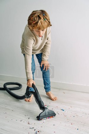Photo for Full body of barefoot boy tidying floor covered with scattered confetti with vacuum cleaner after party - Royalty Free Image