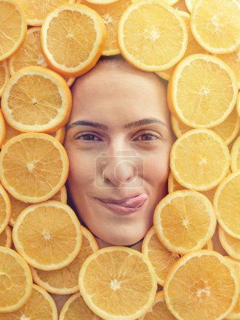 Foto de Modelo femenino satisfecho lamiendo los labios mientras huele las rebanadas de naranja deliciosas maduras dispuestas alrededor de la cara y mirando a la cámara - Imagen libre de derechos