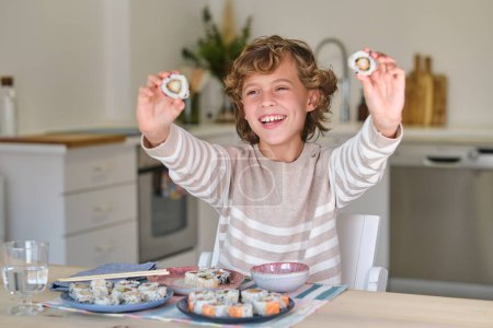 Foto de Niño sonriente divirtiéndose mientras está sentado en la mesa y demostrando rollos de uramaki en la cocina en casa - Imagen libre de derechos