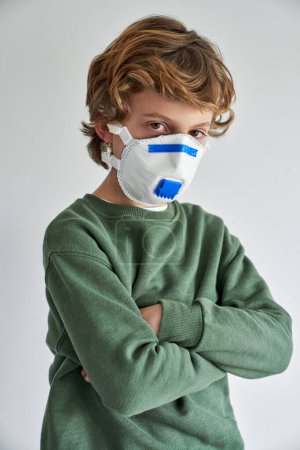 Photo for Preteen child in green sweater and medical respirator with arms crossed self isolating maintaining quarantine and looking at camera - Royalty Free Image