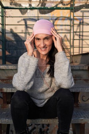Téléchargez les photos : Femme hispanique positive assise sur un escalier en métal et ajustant le symbole de l'écharpe rose du mouvement de santé des femmes et du cancer du sein et regardant la caméra - en image libre de droit