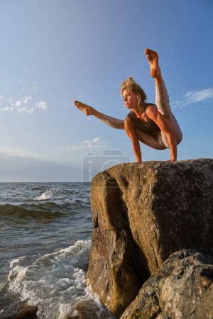 Foto de Desde abajo cuerpo completo de mujer flexible en ropa deportiva balanceándose en brazos mientras practica la pose de luciérnaga en acantilado rocoso durante la sesión de yoga cerca del mar tormentoso - Imagen libre de derechos