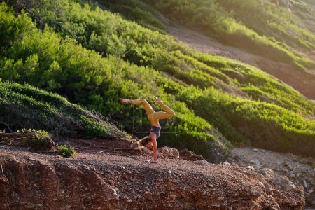 Téléchargez les photos : Vue latérale de la jeune athlète féminine en vêtements de sport prenant Adho Mukha Vrikshasana pose avec les jambes écartées tout en faisant du yoga sur la pente dans les montagnes - en image libre de droit