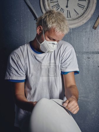 Photo for Focused adult blond haired tattooed male master grinding white handmade surfboard while shaping it with sandpaper in workshop - Royalty Free Image