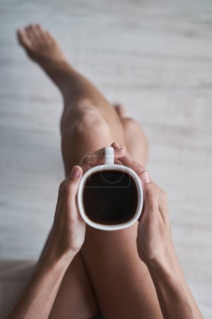 Photo for Overhead view of anonymous female sitting with crossed legs and holding mug of hot aromatic americano during breakfast in morning at home - Royalty Free Image