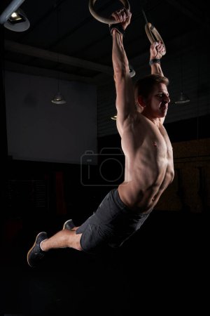 Foto de Vista lateral del cuerpo oscilante atleta musculoso sin camisa en los anillos gimnásticos durante el ejercicio funcional en el gimnasio oscuro - Imagen libre de derechos