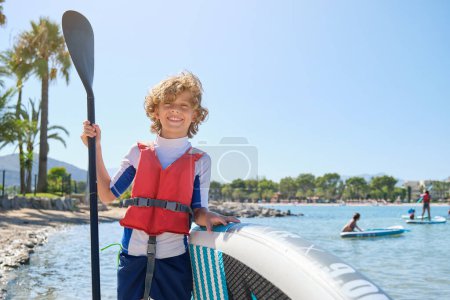Photo for Positive boy in life jacket leaning on paddleboard and smiling while standing on sandy beach with palm trees on sunny summer day - Royalty Free Image