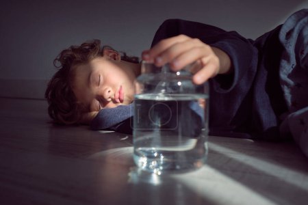 Photo for Ground level of cute little kid with curly hair resting on floor with closed eyes and holding glass transparent jar with clear water in sunbeam - Royalty Free Image