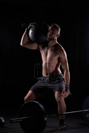 Photo for Powerful muscular male athlete lifting heavy fitness ball on shoulder while doing weightlifting exercise in gym - Royalty Free Image