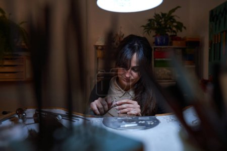 Foto de Orfebre femenino étnico adulto enfocado usando herramienta de grabado a mano mientras crea joyería sentada en el banco de trabajo en la habitación - Imagen libre de derechos