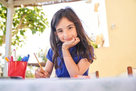 Foto de Sonriente niña hispana con ropa casual mirando a la cámara mientras está sentada en la mesa y dibujando dibujos con plumas coloridas - Imagen libre de derechos