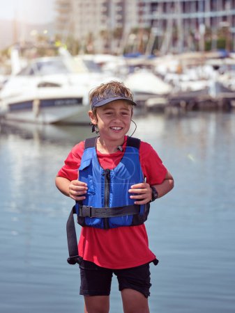 Photo for Positive little boy wearing safety equipment and cap standing near harbor while looking at camera - Royalty Free Image