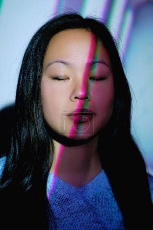 Foto de Encantadora hembra asiática con el pelo largo y negro de pie cerca de la pared con los ojos cerrados en la habitación con luces de neón de colores en la cara - Imagen libre de derechos