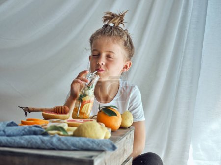 Foto de Niño agradable con los ojos cerrados bebiendo agua saludable con cítricos mientras come en la mesa con frutas sobre fondo blanco en el estudio - Imagen libre de derechos