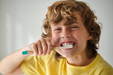 Photo for Child with brown wavy hair in yellow wear brushing teeth with toothbrush while looking at camera - Royalty Free Image