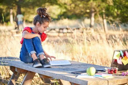 Foto de Cuerpo completo de estudiante preadolescente en ropa casual sentado en la mesa de madera y libro de lectura mientras hace la tarea en el parque en verano - Imagen libre de derechos