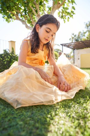 Téléchargez les photos : Pleine longueur de curieux enfant en robe jaune assis sur l'herbe et regardant vers le bas dans la cour arrière verte le week-end en été - en image libre de droit