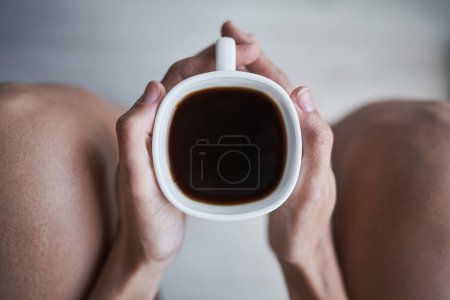 Téléchargez les photos : Vue du dessus de la femelle anonyme tenant une tasse en céramique blanche avec du café noir dans les deux mains pendant qu'elle est assise seule à la maison - en image libre de droit