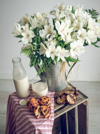 Photo for Bunch of fresh white flowers in metal vase placed near delicious muffins and milk on crate with striped cloth in studio - Royalty Free Image