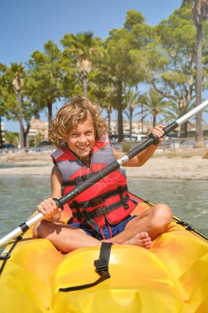 Photo for Cheerful boy with curly hair and life jacket rowing and looking at camera paddling yellow kayak in sea on sunny day - Royalty Free Image