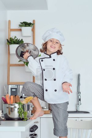 Photo for Positive smiling kid in chef hat and tunic standing near stove with pan lid while cooking in kitchen at home and looking away - Royalty Free Image