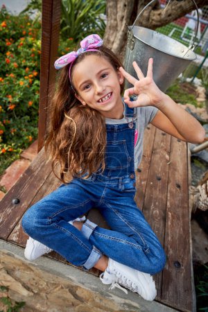 Photo for From above full body of positive girl in overall looking at camera with OK sign while sitting on wooden bench in countryside - Royalty Free Image