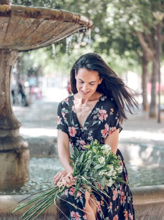 Foto de Feliz mujer encantadora en vestido floral sentado en el borde del borde de piedra de la fuente con ramo de flores en el día de verano en el parque - Imagen libre de derechos