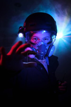 Téléchargez les photos : Enfant en masque à gaz protecteur avec lampe de poche regardant la caméra avec bras tendu tout en cambriolant la menthe avec des néons roses et bleus - en image libre de droit