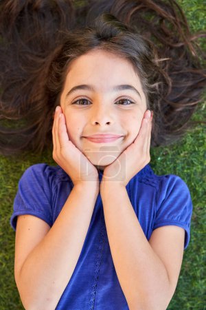 Photo for Top view of positive child with long hair touching face and looking at camera while lying on green lawn on weekend summer day - Royalty Free Image